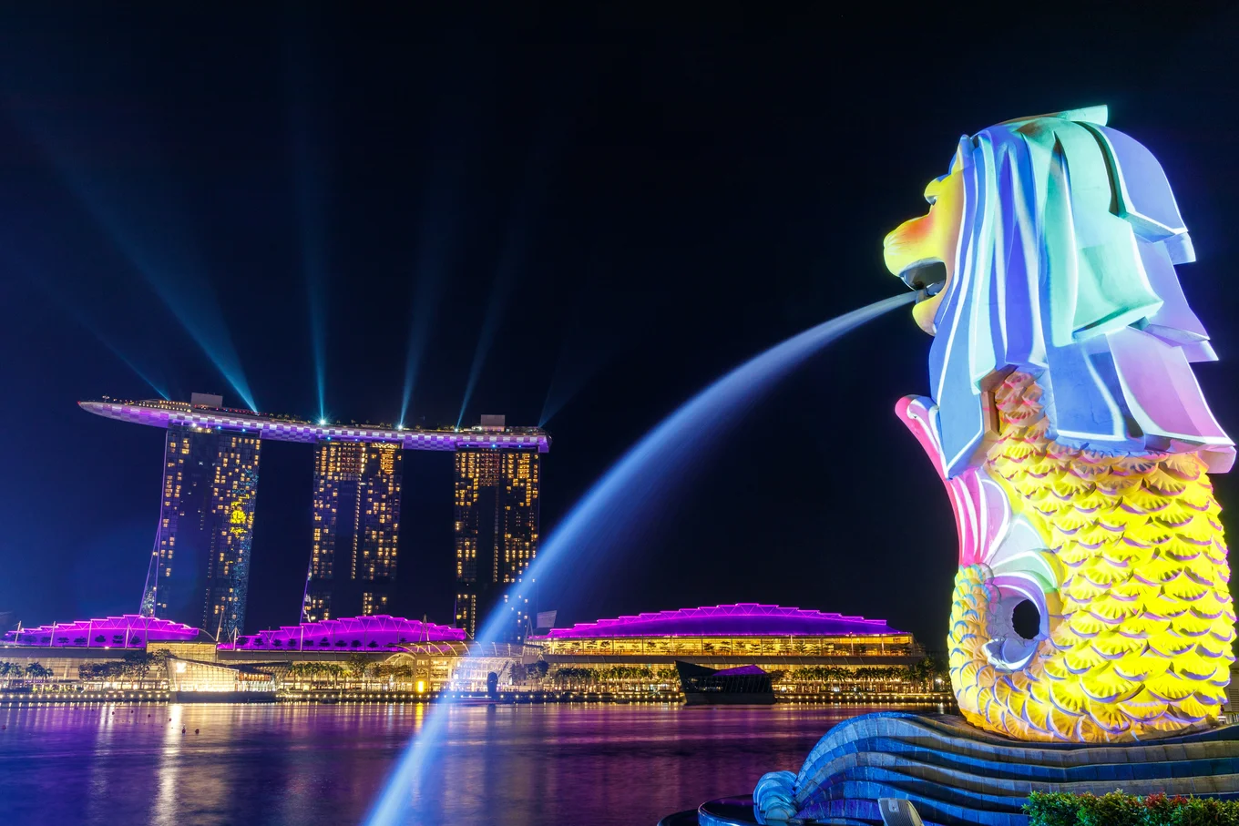 Singapore at night in multicoloured lighting, showcasing the Marina Bay Sands Hotel and one of the Merlion statues.